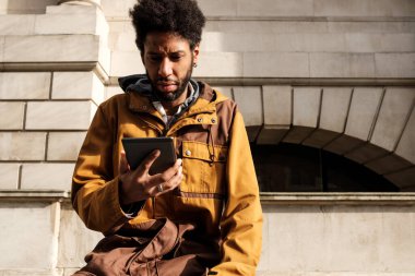Taş bir binanın önünde oturan afro saçlı genç adam güneşli bir günde şık sarı ve kahverengi bir ceket giyerken dijital tabletle meşgul oluyor.