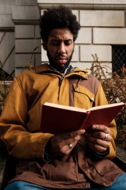 Afro saç stili olan odaklanmış genç siyah bir adam bankta oturuyor, açık havada şık bir kahverengi ve sarı ceket giyerken kırmızı bir kitap okumaya dalmış.