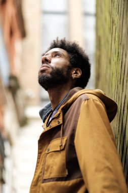 Şık sakallı, afro saçlı, modaya uygun kahverengi ceketli ve küpeli dar bir şehir sokağında özenle dik dik bakan genç bir zenci portresi.