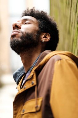 Afro saçlı ve küpeli genç adam yosunlu bir duvara yaslanıyor, gözlerini kapıyor ve derin derin nefes alıyor, huzur ve sükunetin tadını çıkarıyor.