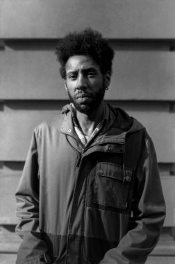 Tahıllı tek renkli analog fotoğraf kapüşonlu ceketli genç bir adamı, desenli kıvrımlı demir bir arka planda, zamansız otantiklik yayarken yakalıyor.