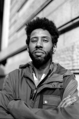 Kentin bulanık arka planında duran, katlanmış kolları ve sakalı olan özgüvenli Afro-Amerikan bir adamın 35 mm 'lik tek renkli portresi.