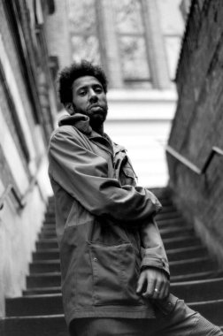 Kolları çapraz bir şekilde merdivenlerde duran, ceket giyen ve ciddi görünen genç bir adamın tek renkli, analog bir portresi.