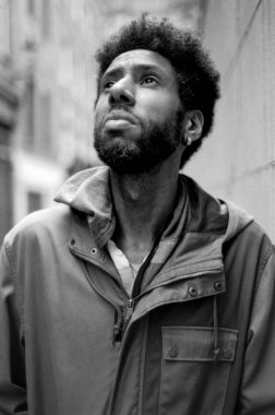 Afro saçlı, kapüşonlu ceketli, şehir ortamında umut verici bir ifadeyle yukarı bakan genç bir adamı gösteren siyah beyaz bir fotoğraf.