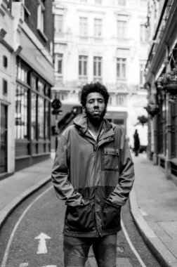Londra 'da dar bir caddenin ortasında elleri cebinde kapüşonlu ceket ve kot pantolon giymiş genç bir adamı gösteren tek renkli, klasik bir fotoğraf.