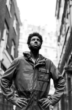 Binalar arasında dar bir sokakta duran, elleri kalçasında yukarı bakan kendine güvenen bir turistin tahıllı siyah beyaz analog fotoğrafı.