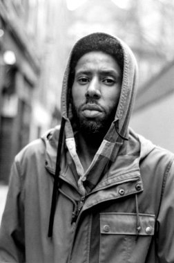 Taneli monokrom analog görüntü kapüşonlu ceketli genç bir adam görüntüsü, güvenli bir şekilde kameraya bakıyor, bulanık bir şehir zeminine karşı, zamansız bir sokak titreşimini yakalıyor