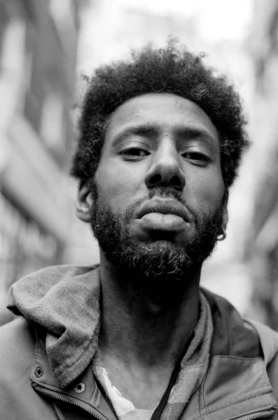 Kapüşonlu ceketli genç bir adamın monokrom analog film portresi, şehir arkaplanının ortasında duruyor, cesur, otantik bir ruh hali yakalayan ciddi bir ifade yayıyor.