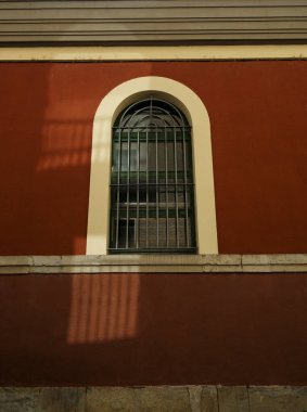 Kahverengi bina duvarı koyu güvenlik parmaklıklarıyla donatılmış kemerli bir pencere sunuyor. Sıcak güneş ışığı alıyor ve desenli ön cepheye karşı desenli bir gölge oluşturuyor.