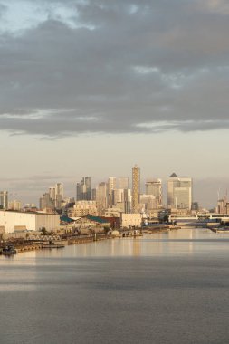 Kanarya Rıhtımı gökdelenleri ve sakin nehir kıyısındaki ticari binaları gösteriyor. Bulutlu bir gökyüzünün altında, gün doğumunda yansıtan bir ışık yaratıyor.