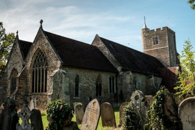 Gotik mimari ve çan kulesinin yer aldığı tarihi taş kilise, mavi gökyüzüne karşı dikilmiş, eski çağlardan kalma mezar taşları ve geleneksel bir kilise bahçesindeki yeşil çimlerle çevrili.