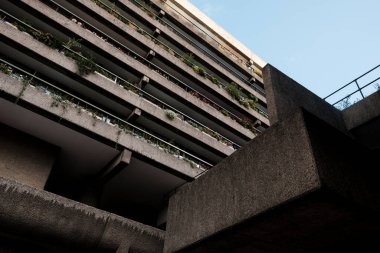 Barbika mülkünün beton bloğu alçak açıdan görülüyor, saksı bitkileri ve korkuluklarla dolu balkonlar, berrak gökyüzüne karşı geometrik vahşet aralığı.