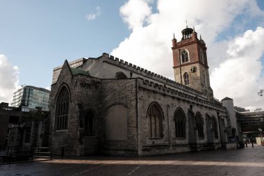 Aziz Giles 'ın kötürüm olmayan kilisesi dimdik ayakta duruyor. Bulutlu bir gökyüzünün altındaki modern barbika mimarisi ile çelişen saatli taş bir kulenin yer aldığı tarihi bir yer.
