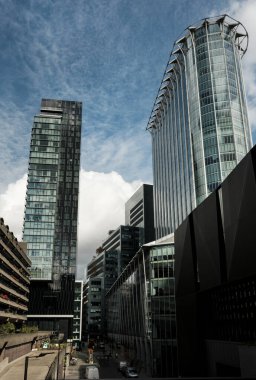 Londra şehir mimarisi, parlak bulutlu gökyüzüne karşı yükselen cam cepheli modern gökdelenleri sergiliyor, dinamik çizgiler ve geometrik desenlerle kentsel bir manzara yaratıyor.