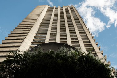 Barbika emlak binası, beton dokusu ve geometrik çizgileri açık mavi gökyüzünün altında beyaz bulutlarla uzanan vahşi mimari kompozisyonu sergiliyor.