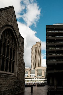 Beyaz bulutlu mavi gökyüzüne yükselen acımasız beton gökdelen, ön planda yeşil yapraklarla birlikte Londra 'nın eşsiz barbika arazisinin tasarımını sergiliyor.