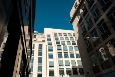 Geometrik cephelerin ve cam pencerelerin modern şehir ufuk çizgisi, mimari desenleri vurgulayan alçak açı görünümü, güçlü gölgeler ve berrak mavi gökyüzü altında yansıtıcı yüzeyler