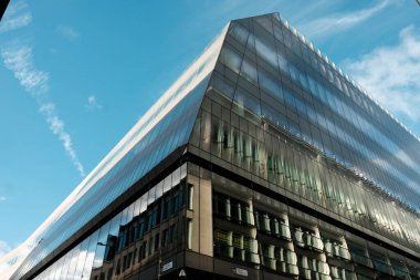 Çağdaş cam gökdelen cephesi, açık mavi gökyüzünü ve geleneksel bir binayı yansıtıyor, şehir ortamında mimari karşıtlıklarla dinamik bir şehir manzarası sunuyor.