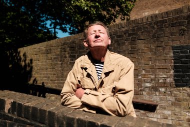 Yaşlı adam gözleri kapalı ve kollarını kavuşturmuş, sıcak güneş ışığını eskimiş tuğla bir duvara dayayıp derin bir huzur, memnuniyet ve sükunet hissi yayıyor.