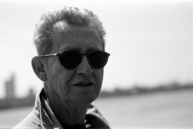 Yaşlı bir adamın güneş gözlüğü taktığı, açık havada güneşli bir günün tadını çıkardığı, arka planda bulanık bir su birikintisi ve şehir manzarası olduğunu gösteren tek renkli, tek renkli, klasik bir fotoğraf.