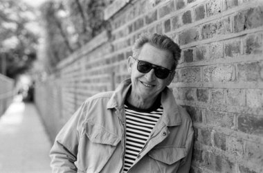 Güneş gözlüğü takan ve tuğladan bir duvara yaslanan gülümseyen yaşlı bir adamın tek renkli, analog film portresi gerçek bir mutluluk anı ve zamansız bir stil yakalıyor.