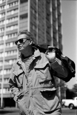 Güneş gözlüklü ve açık renk ceketli yaşlı bir adamın arka planda büyük bir konut binası bulunan bir çanta taşıdığı tek renkli 35 mm 'lik bir fotoğraf.