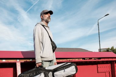 Kıdemli müzisyen, güneşli bir günde kırmızı bir köprüde enstrüman çantasını taşıyor, düşünceli bir ifadeyle mesafeye bakıyor.