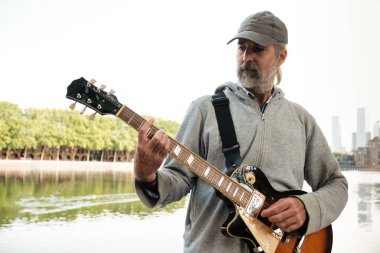 Beyzbol şapkası takan yaşlı sokak müzisyeni nehir kenarında elektro gitar çalıyor ve kanarya iskelesinin silueti Londra 'da çarpıcı bir zemin sağlıyor.