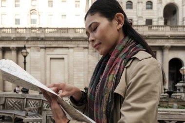 Trençkotlu ve renkli eşarplı bir kadın kağıt harita okuyor, tarihi bir Avrupa şehrini geziyor ve keşfediyor, yeni bir yerde yol gösteriyor.