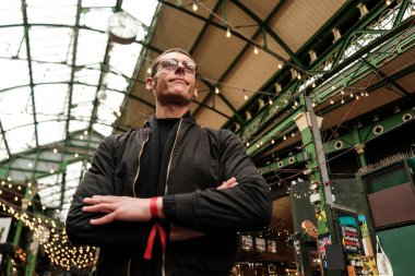 Düşük açılı, çapraz kollu ve kırmızı bileklikli genç bir adam canlı kasaba pazarında duruyor. Güney Afrika, Londra 'da bulunan tarihi bir gıda pazarı.