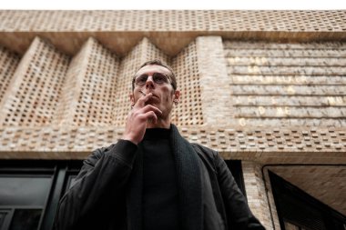 Modern tuğlalı bir binanın önünde sigara içen şık bir adamın düşük açılı görüntüsü büyüleyici bir şehir manzarası yaratıyor.