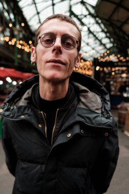 Gözlüklü ve kışlık giysili kendinden emin genç adam şehir pazarında duruyor, Londra 'da, arka planda bulanık ışıklar ve market tezgahları var.