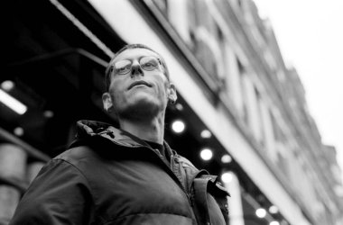 Arkaplanda bulanık bir binayla şehir ortamında gözlük takan dalgın bir adamın tek renkli analog 35mm 'lik fotoğrafı.