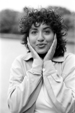 Tahıllı monokrom analog fotoğraf kıvırcık saçlı ve burun piercingli genç bir kadın, elleriyle yüzünü tutuyor, zarif ve anlamlı bir portre yaratıyor.