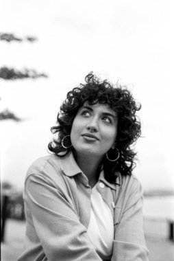 Düşünceli, kıvırcık saçlı, burun piercingli, açık renk ceketli, büyük halka küpeli, umutla dik dik bakan genç bir kadının tek renkli portresi.