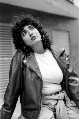 Kumlu siyah beyaz analog fotoğraf şık bir deri ceket ve halka küpe takan 35 mm 'lik filmle çekilen düşünceli bir ifadeyle yukarıya bakan genç bir kadını çekiyor.