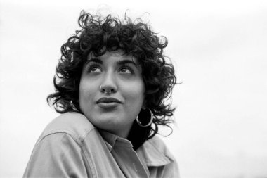 Tahıllı monokrom analog fotoğraf kıvırcık saçlı, burun piercingli, halka küpeli, temiz gökyüzüne dikkatlice bakan genç bir kadını yakalıyor.