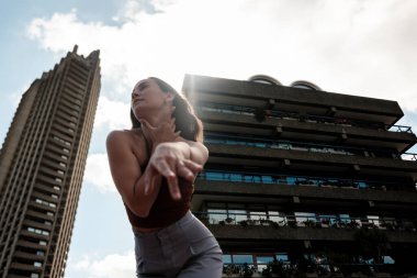Genç bayan dansçı, Londra 'daki barbican binalarının önünde modern bir dans koreografisi sergiliyor. Güneş ışığı bulutların arasından süzülüyor.