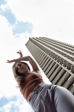 Dansçı zarif bir pozla modern bir gökdelenin arka planına doğru yükseliyor. Hareketleri yukarıdaki bulutlu gökyüzünü çeken alçak açıyla vurgulanıyor.