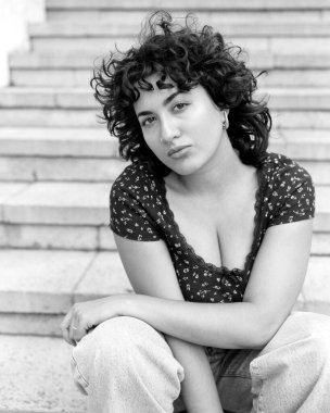 Grady monokrom orta format film portresi kıvırcık saçlı ve burun piercingli, basamaklarda oturan, özgüven ve bireysellik duygusu yayan şık bir genç kadın.