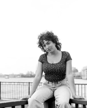 Taneli monokrom orta formatlı bir film portresi. Nehir kenarındaki korkulukta oturan, rahat bir kot ve çiçekli bir bluz giymiş, sakin ve derin düşüncelere dalmış genç bir kadını çekiyor.