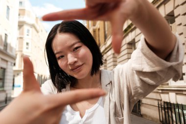 Genç kadın parmaklarıyla bir çerçeve yaratıyor, gençlik enerjisini mimari zemin ile harmanlayan dinamik bir kompozisyonda kentsel manzarayı neşeyle yakalıyor.