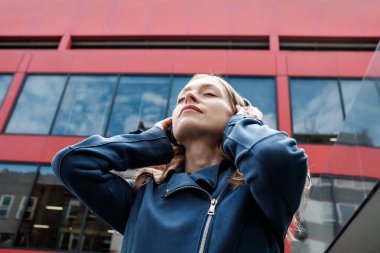 Kulaklık takan genç bir kadın, gözleri kapalı ve elleri kulaklarında müzik dinliyor. Şehir ortamında modern kırmızı bir binanın önünde duruyor.