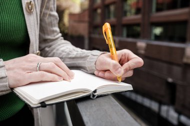 Bir kadının ellerinin turuncu kalemle deftere yazarken, potansiyel olarak fikirleri karalarken, bir yapılacaklar listesi yaratırken ya da açık şehir ortamında düşüncelerini günlüğe yazarken yakın plan.