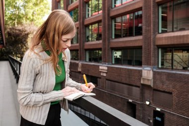 İş kadını, modern bir ofis binasının balkonunda çalışırken küçük bir not defterine not alıyor. Çalışma saatleri boyunca fikir ve gözlemleri yakalıyor.