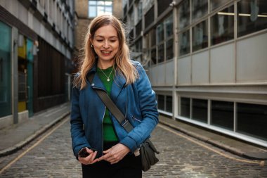 Londra 'da büyüleyici bir kaldırım taşı sokakta yürüyen mutlu bir iş kadını, elinde akıllı bir telefonla yeşil bir kazağın üzerine şık bir mavi ceket giyer, kendine güven ve neşe saçar.
