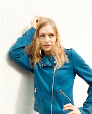 Mavi ceketli sarışın kadın beyaz bir duvara yaslanıyor, saçıyla oynuyor ve başka tarafa bakıyor, analog orta formatlı film kamerasıyla çekiliyor, film tahılları ve klasik renkler yer alıyor.