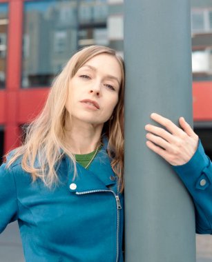 Deniz mavisi ceketli sarışın kadın şehir ortamında gri bir direğe yaslanıyor. Düşüncelere dalmış, sakin bir hava yayıyor.