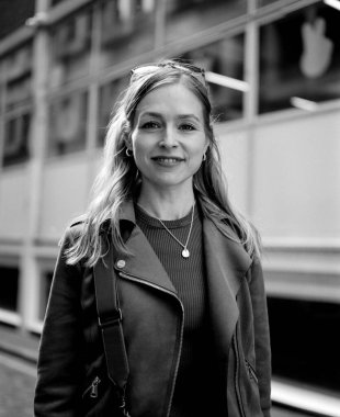 Deri ceket ve güneş gözlüğü takan gülümseyen bir kadının tek renkli orta formatlı film portresi, kendine güvenen ve şık bir tavırla modern bir şehir ortamında duruyor.