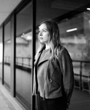 İş kadını şık giyinmiş, düşünceli bir şekilde büyük bir ofis penceresinden dışarı bakıyor, tek renkli, tek renkli bir film formatında çalışmalarını yansıtıyor.